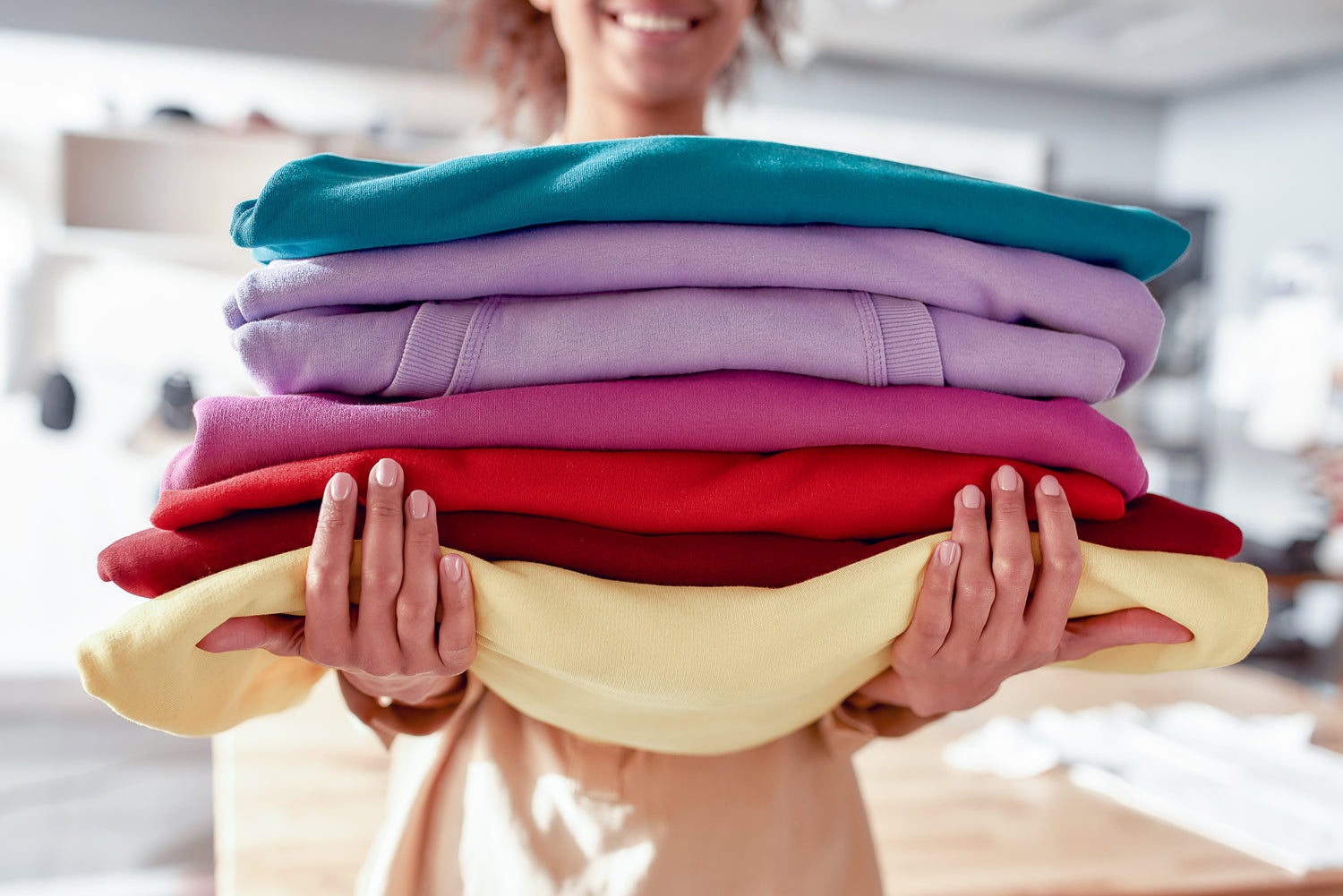 woman holding 8 folded sweatshirts of different colors